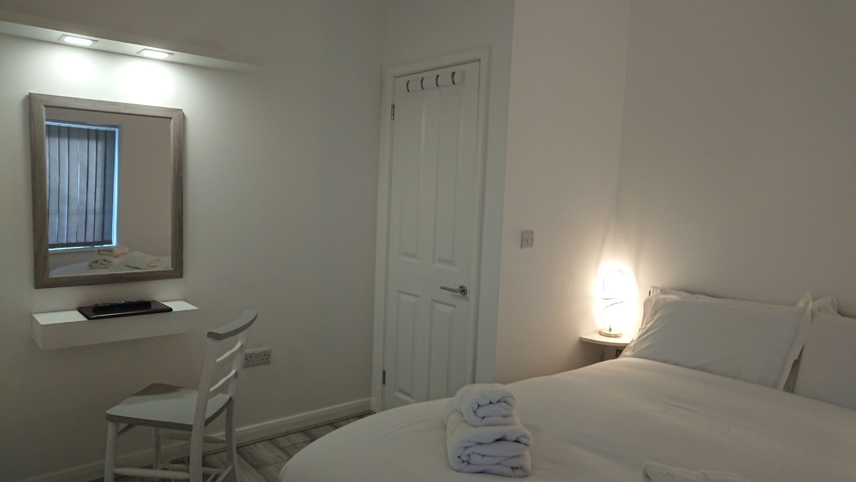 Habitación de hotel con cama, escritorio con silla, espejo, puerta y lámpara de noche.