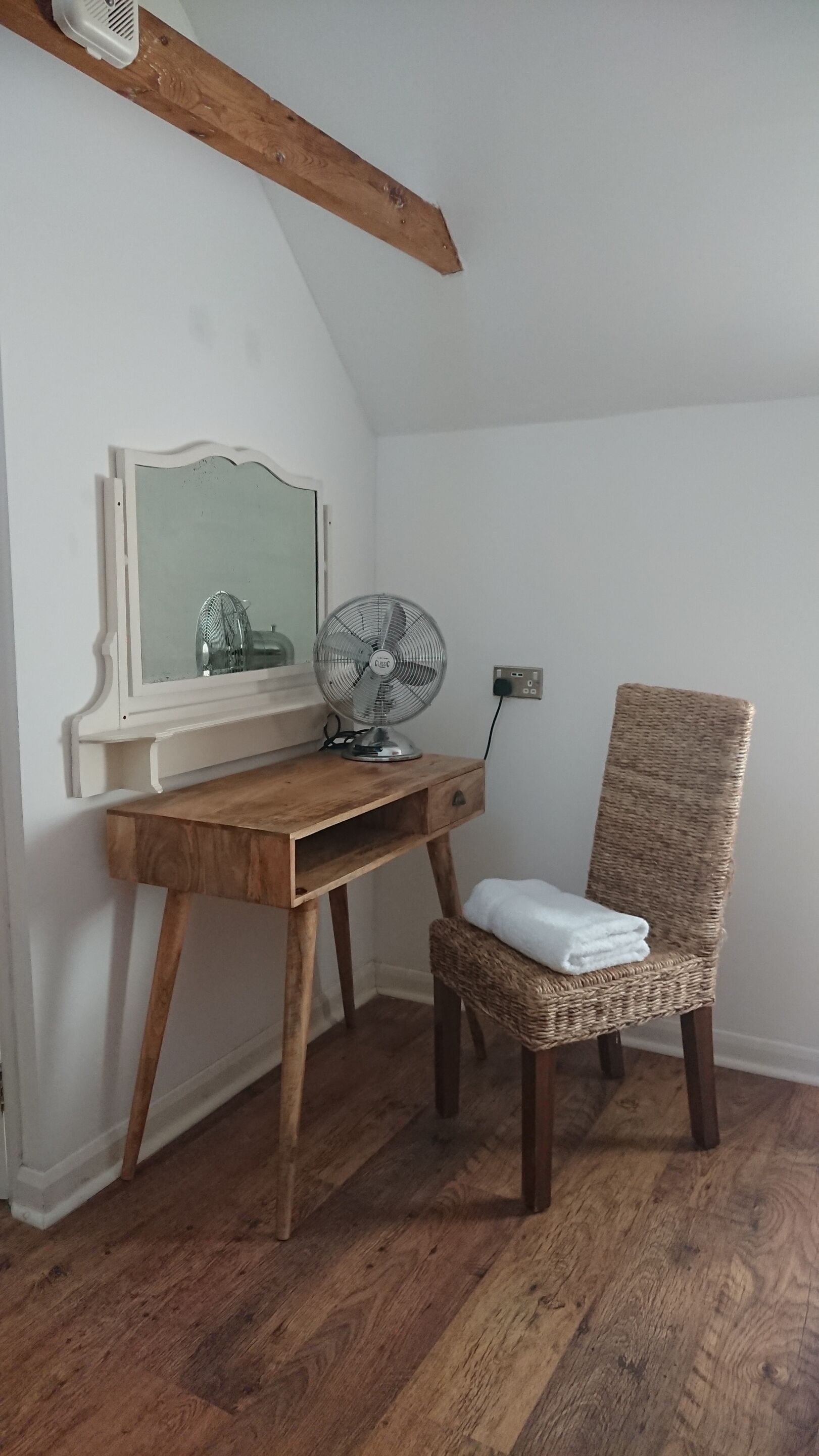 A room with a wooden desk, a wicker chair, a standing fan, and a mirror.