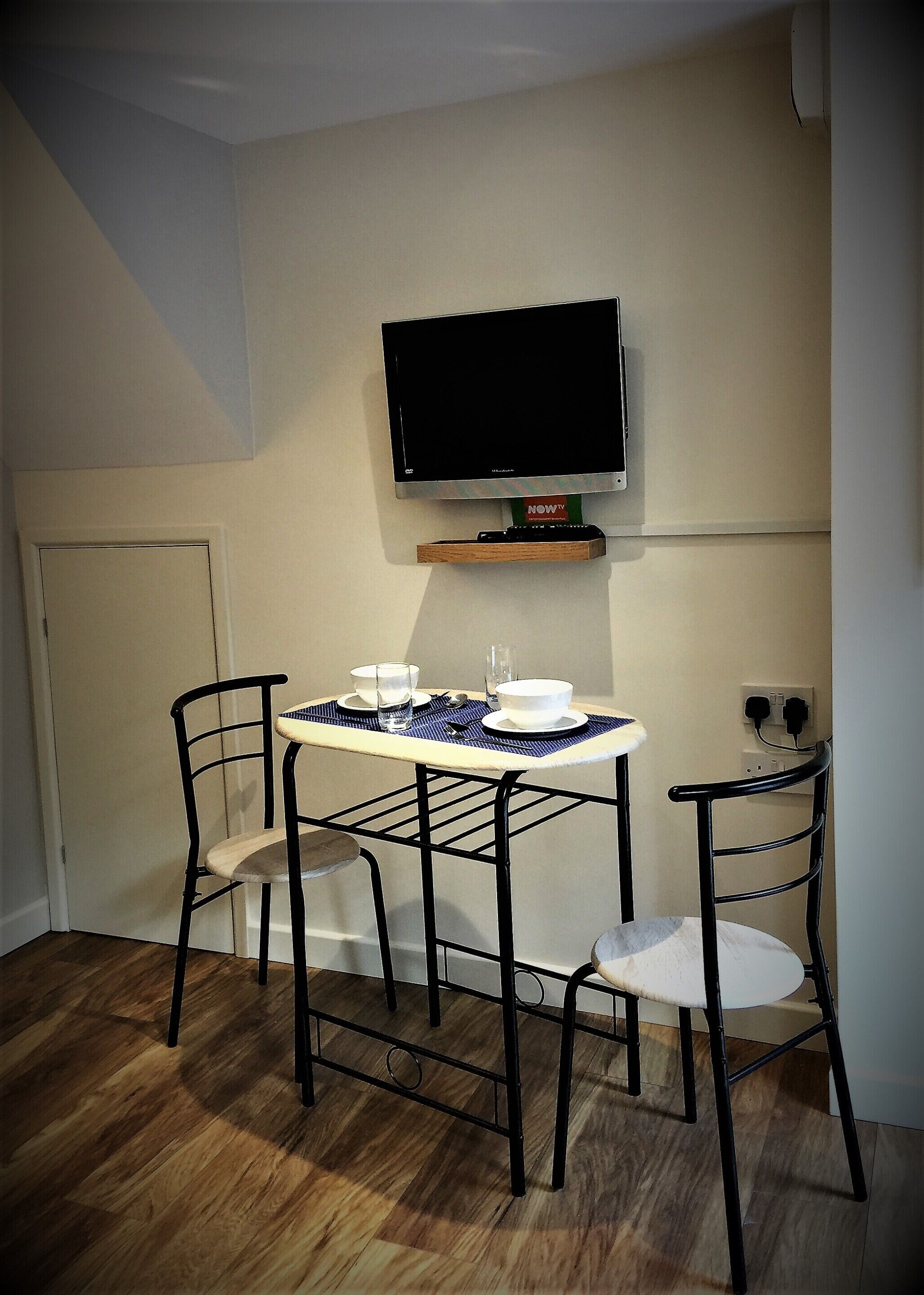 A small dining area with a table and two chairs, a television mounted on the wall, and a door in the background.