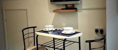 A small dining area with a table and two chairs, a television mounted on the wall, and a door in the background.