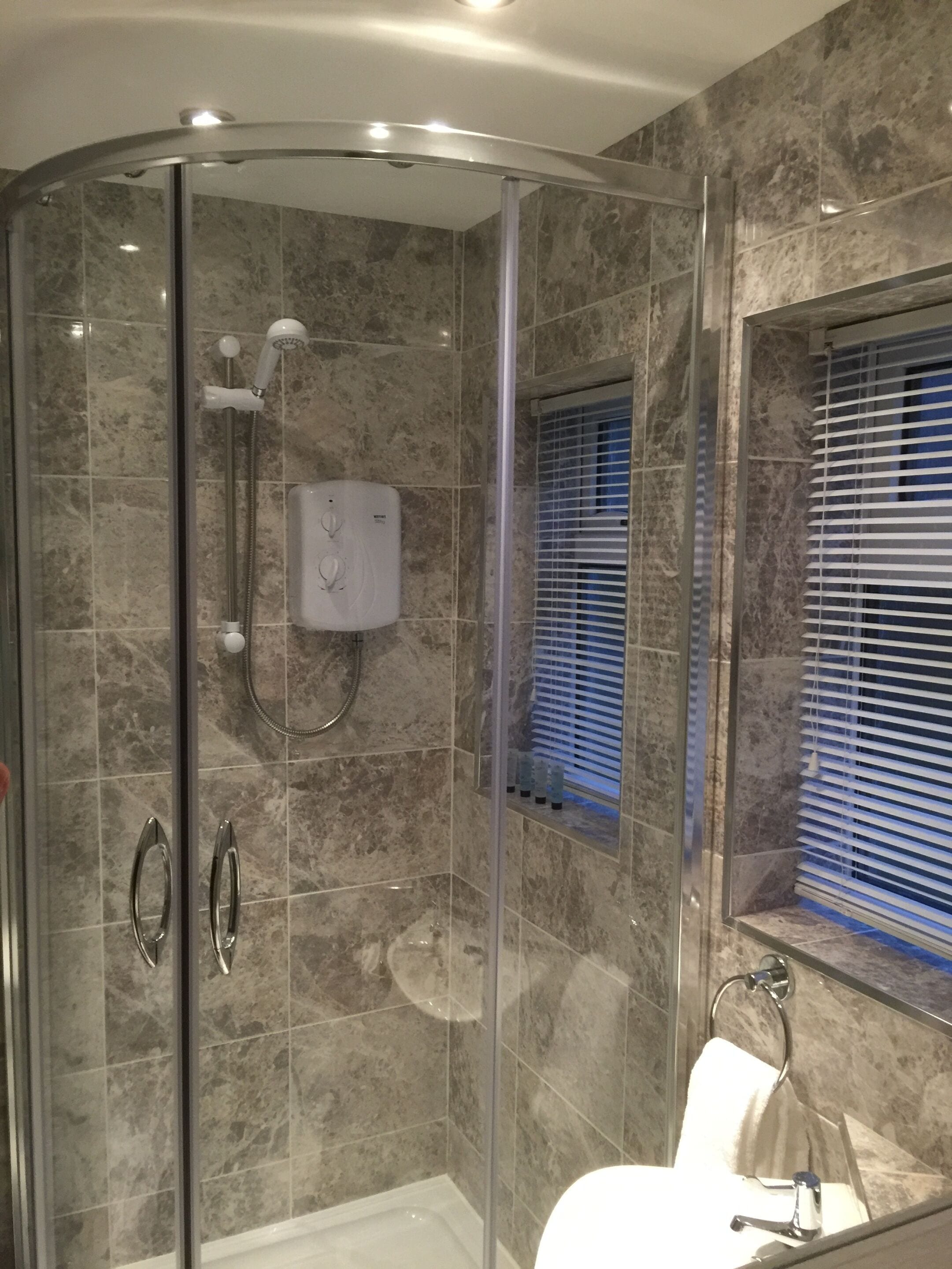 A modern bathroom with a glass-enclosed shower, a marble-tiled wall, a built-in sink, and a window with blinds.