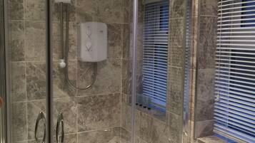 A modern bathroom with a glass-enclosed shower, a marble-tiled wall, a built-in sink, and a window with blinds.