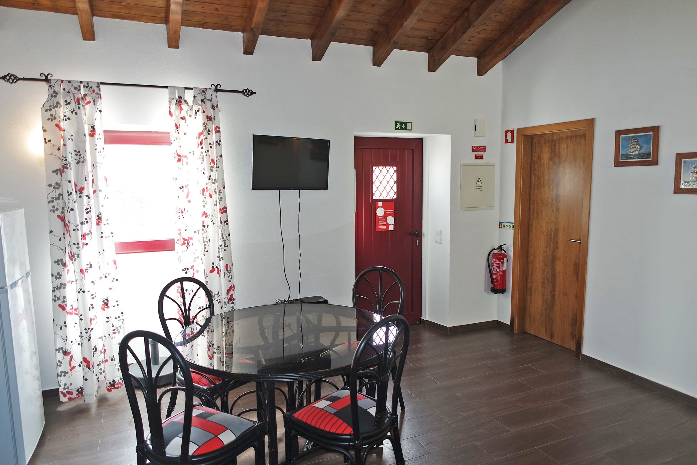 A dining area with a round glass table, black chairs with red cushions, a television on the wall, a red door, and a fire extinguisher mounted on the wall.