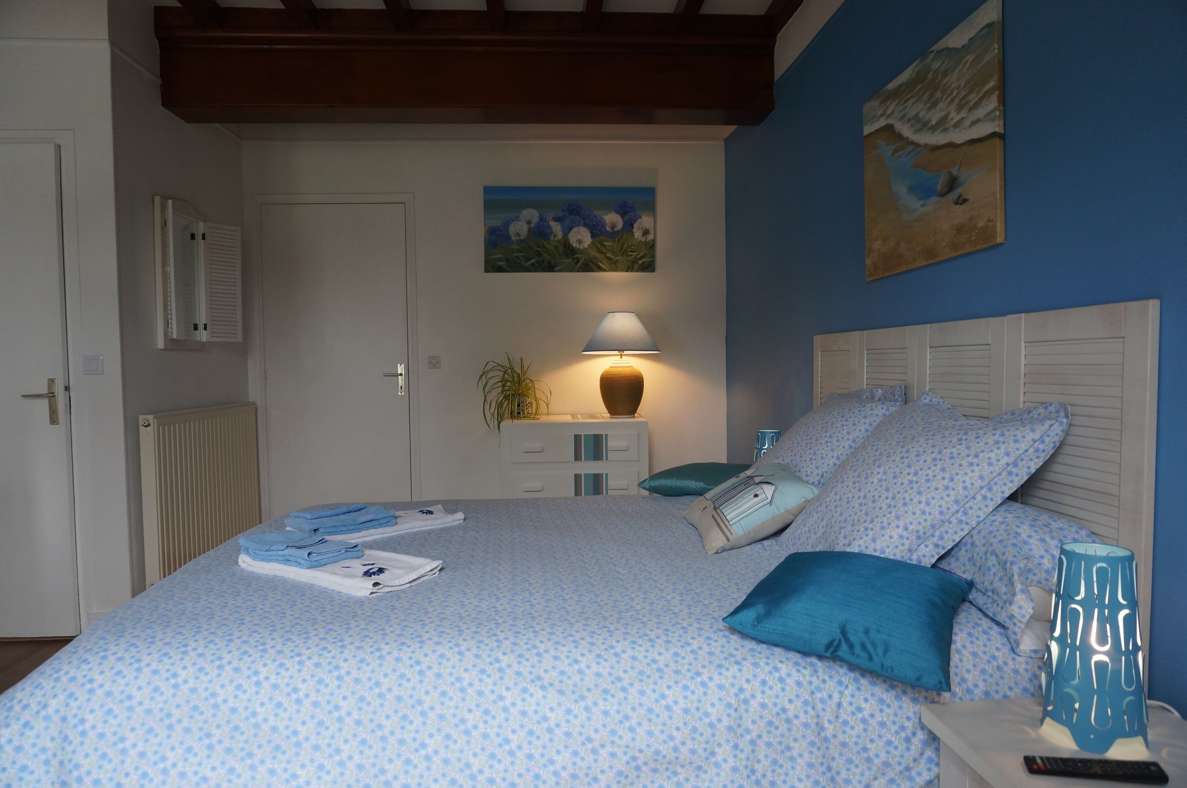 Un dormitorio con cama, almohadas, mesita de noche, lámpara y un cuadro en la pared.