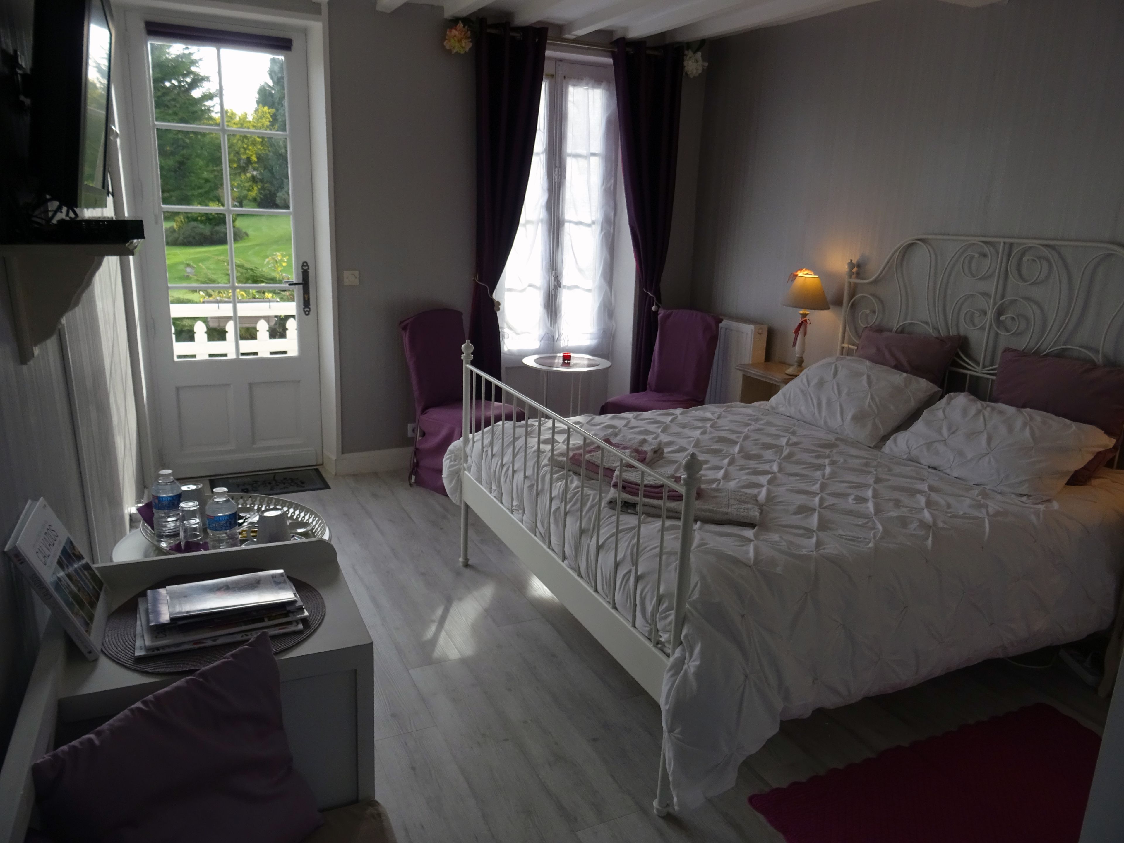 Ein Schlafzimmer mit einem weißen Bett, violetten Stühlen, einem kleinen Tisch und einem Fenster mit Vorhängen.