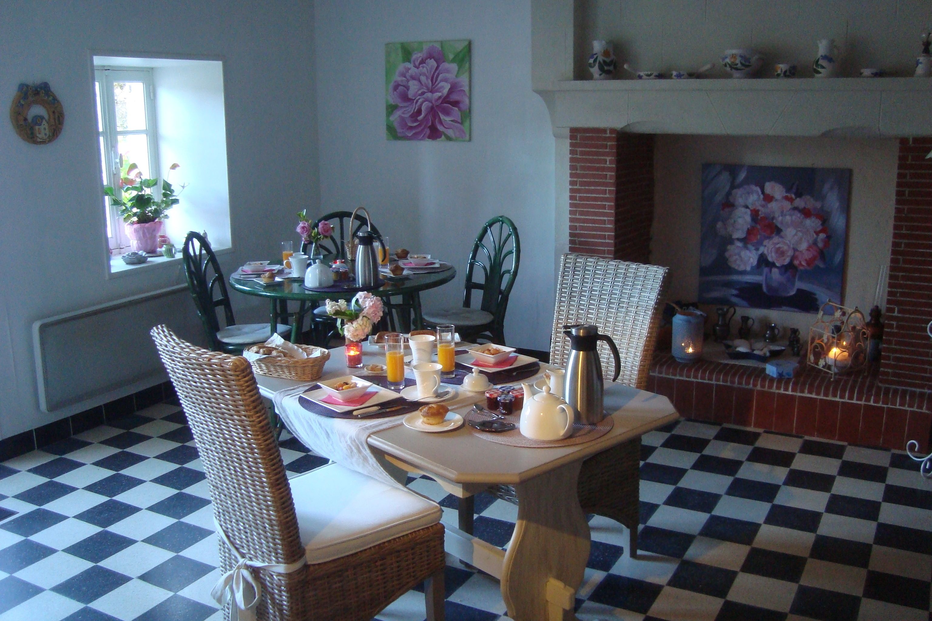 Een eetruimte met een zwart-wit schachbordvloer, een ontbijttafel, rieten stoelen en een open haard met daarboven een schilderij.