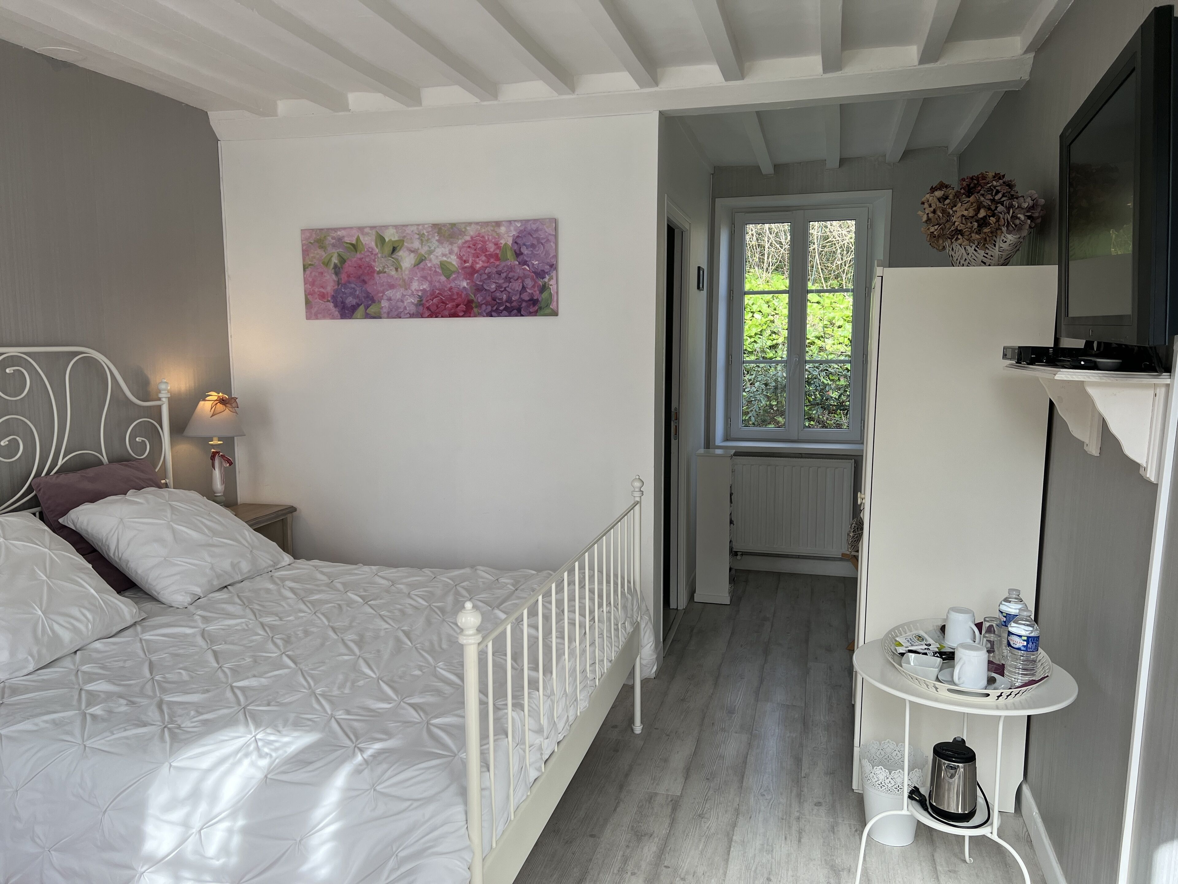 Une chambre à coucher avec un lit, une table de chevet avec une lampe, un tableau au mur, une fenêtre et une petite table avec une bouilloire et des tasses.