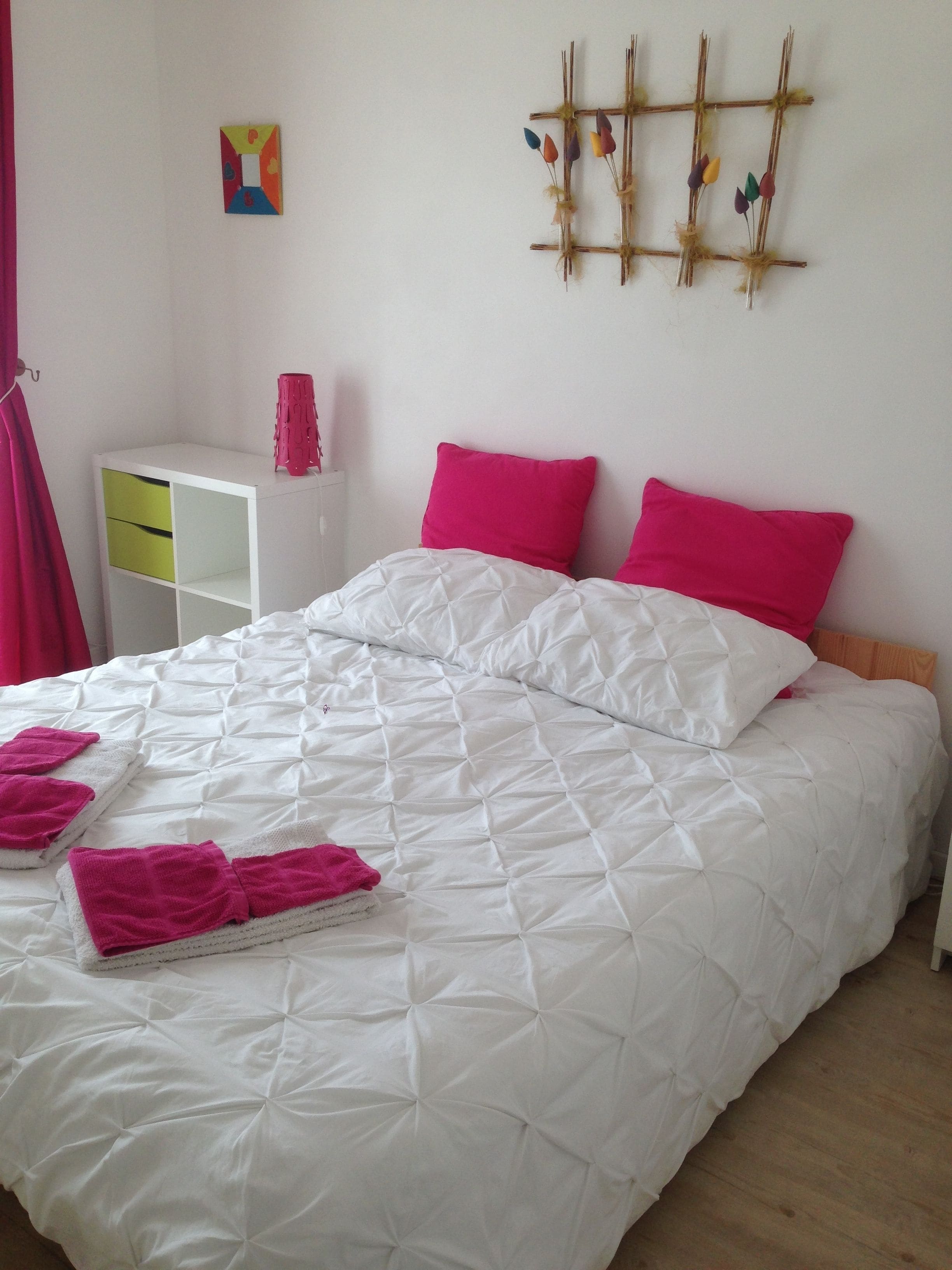 Een net opgemaakt bed met wit en roze beddengoed, een klein wit nachtkastje met een roze lamp, en een decoratie aan de muur.