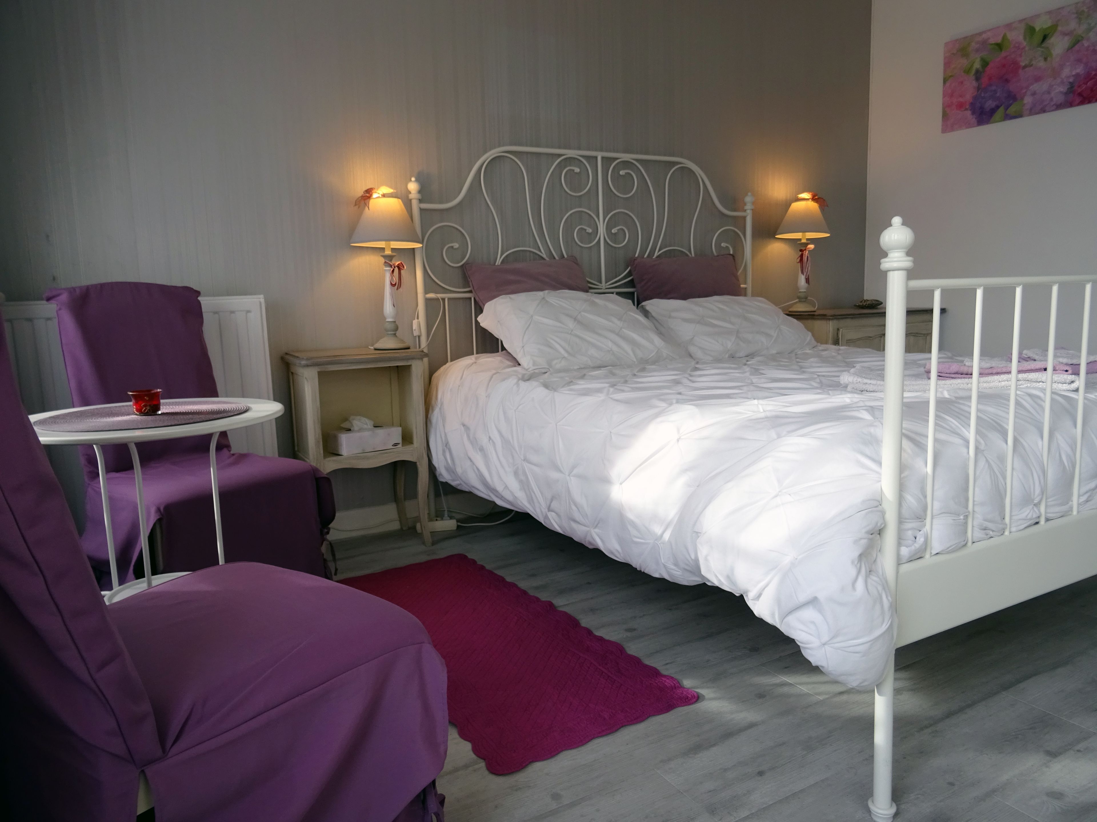 Une chambre à coucher avec un lit en métal blanc, des chaises violettes, une petite table ronde et deux lampes de chevet.