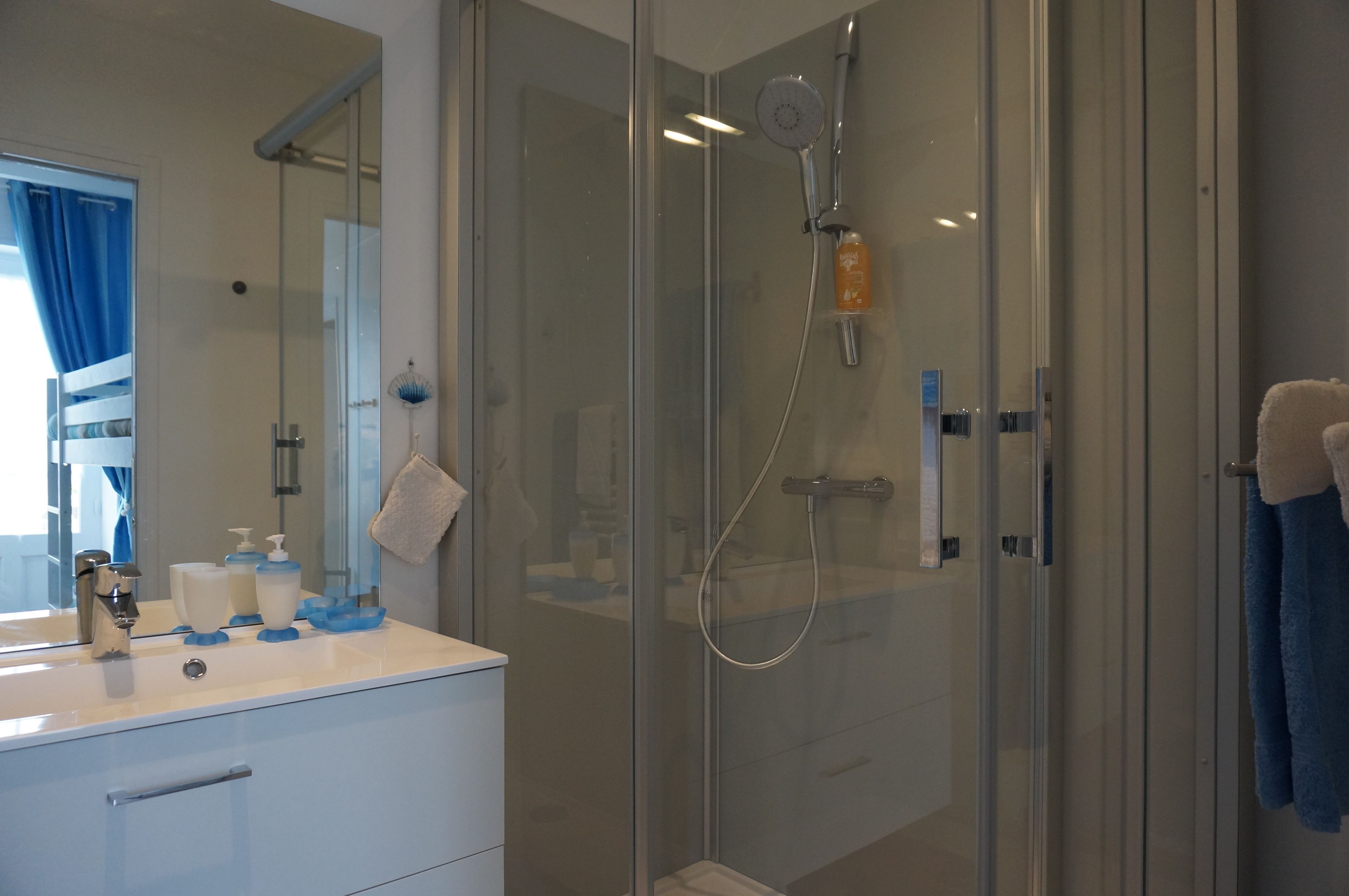 Een moderne badkamer met een douchecabine van glas, een witte wastafel met blauwe zeepdispensers, en een spiegel met een blauw gordijn.