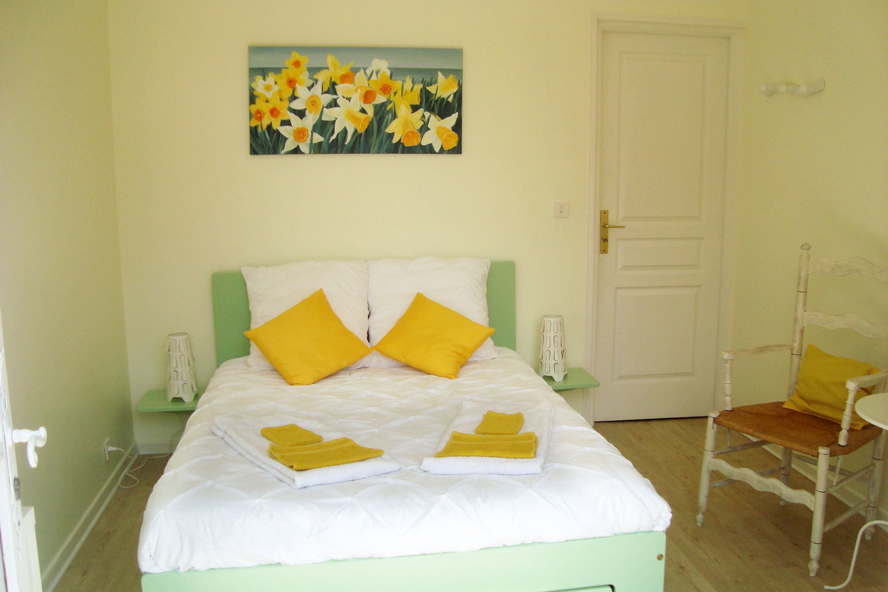 Een bed met witte beddengoed en twee gele kussens, een schilderij van gele bloemen aan de muur, en een houten stoel met een geel kussen.
