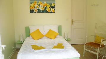 Een bed met witte beddengoed en twee gele kussens, een schilderij van gele bloemen aan de muur, en een houten stoel met een geel kussen.