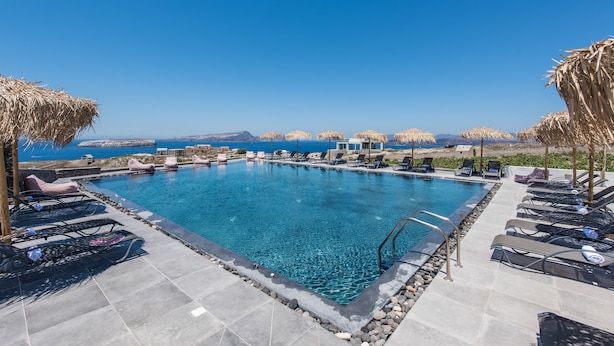 Une piscine à l’eau d’un bleu cristallin, entourée de chaises longues et de parasols, offrant une vue sur la mer et les collines lointaines.