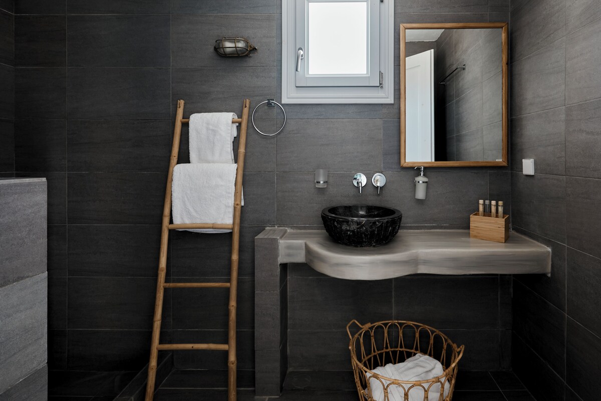 A modern bathroom with dark tiles, a wooden ladder, a white towel, a black sink, and a mirror.