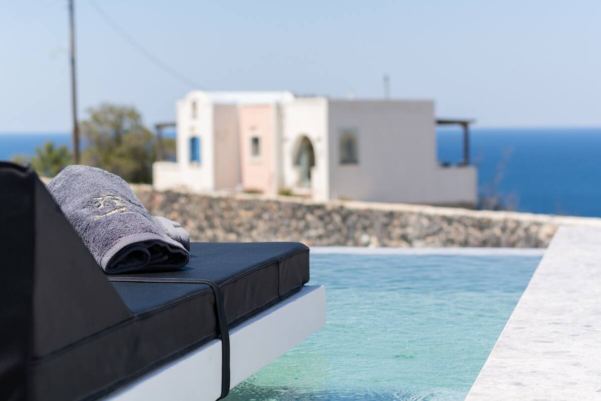 Zona de piscina amb una cadira relaxant i una tovallola, pared de pedra i un edifici amb vistes al mar al fons.