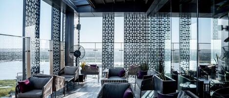 Un espacio interior moderno con ventanas amplias, vista a un cuerpo de agua y una zona de estar con sillas y una mesa.