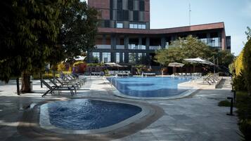 Zona de piscina con dos piscinas, tumbonas y sombrillas. Se ve un edificio con ventanas grandes al fondo.