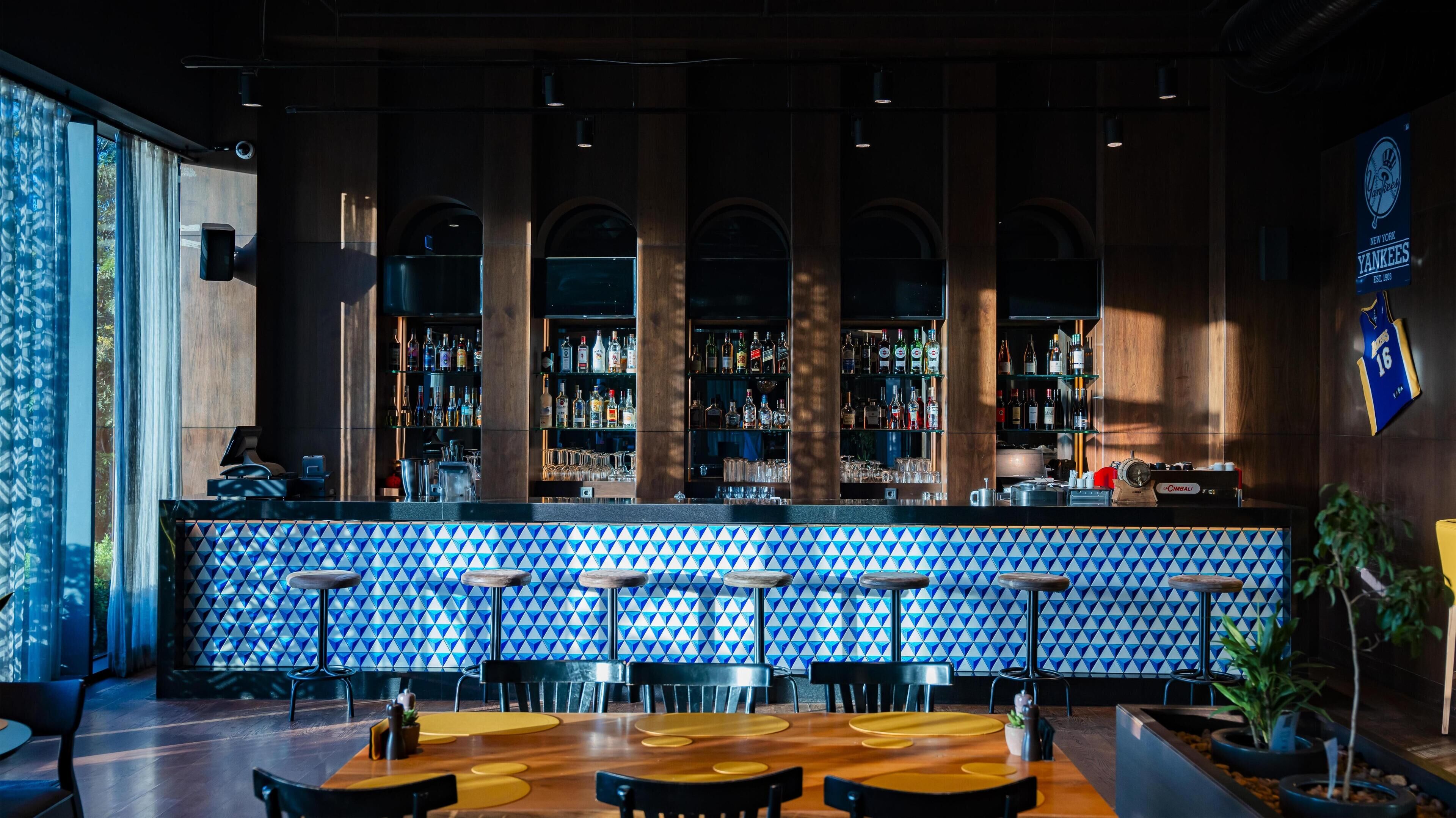 Un bar con pared de azulejos blancos y azules, mostrador de madera y varias zonas de estar con sillas negras.