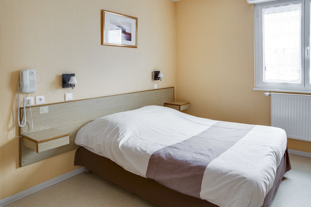 Habitación de hotel con cama, mesitas de noche, teléfono, lámpara, un cuadro enmarcado y una ventana con persianas.
