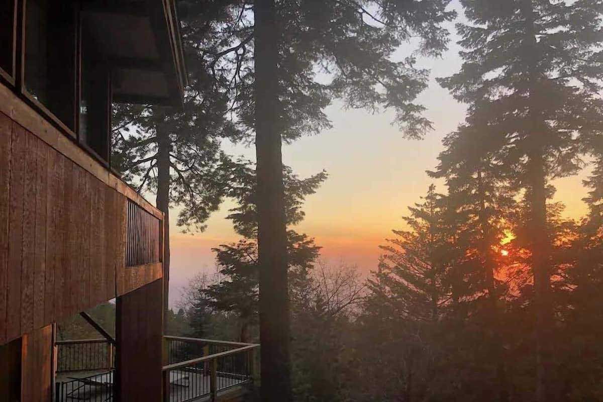 A wooden cabin with a balcony overlooking a forested area during sunset.