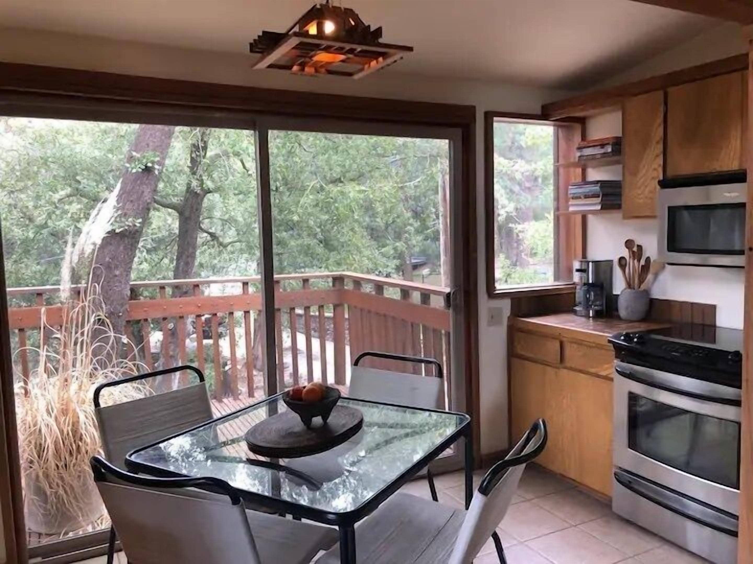 Una cocina moderna con una mesa y sillas de vidrio, armarios de madera y vistas a árboles a través de grandes ventanales.