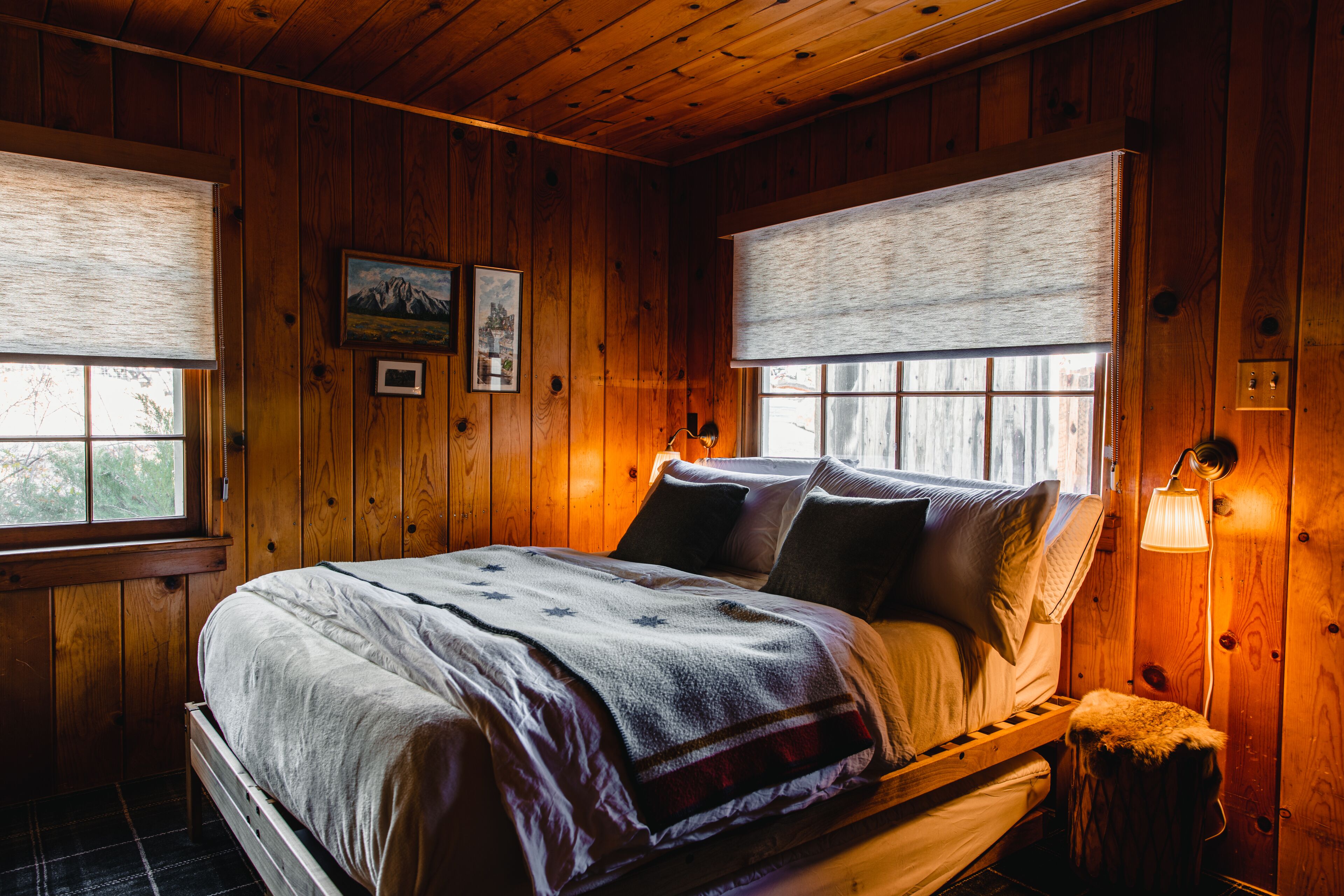 Un dormitorio de cabaña de madera con una cama, dos ventanas con persianas, una mesita de noche y una lámpara fijada a la pared.