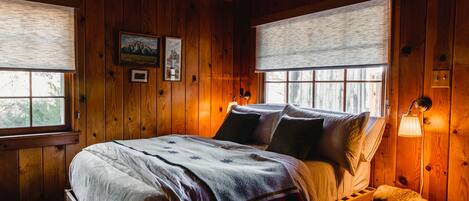 Un dormitorio de cabaña de madera con una cama, dos ventanas con persianas, una mesita de noche y una lámpara fijada a la pared.
