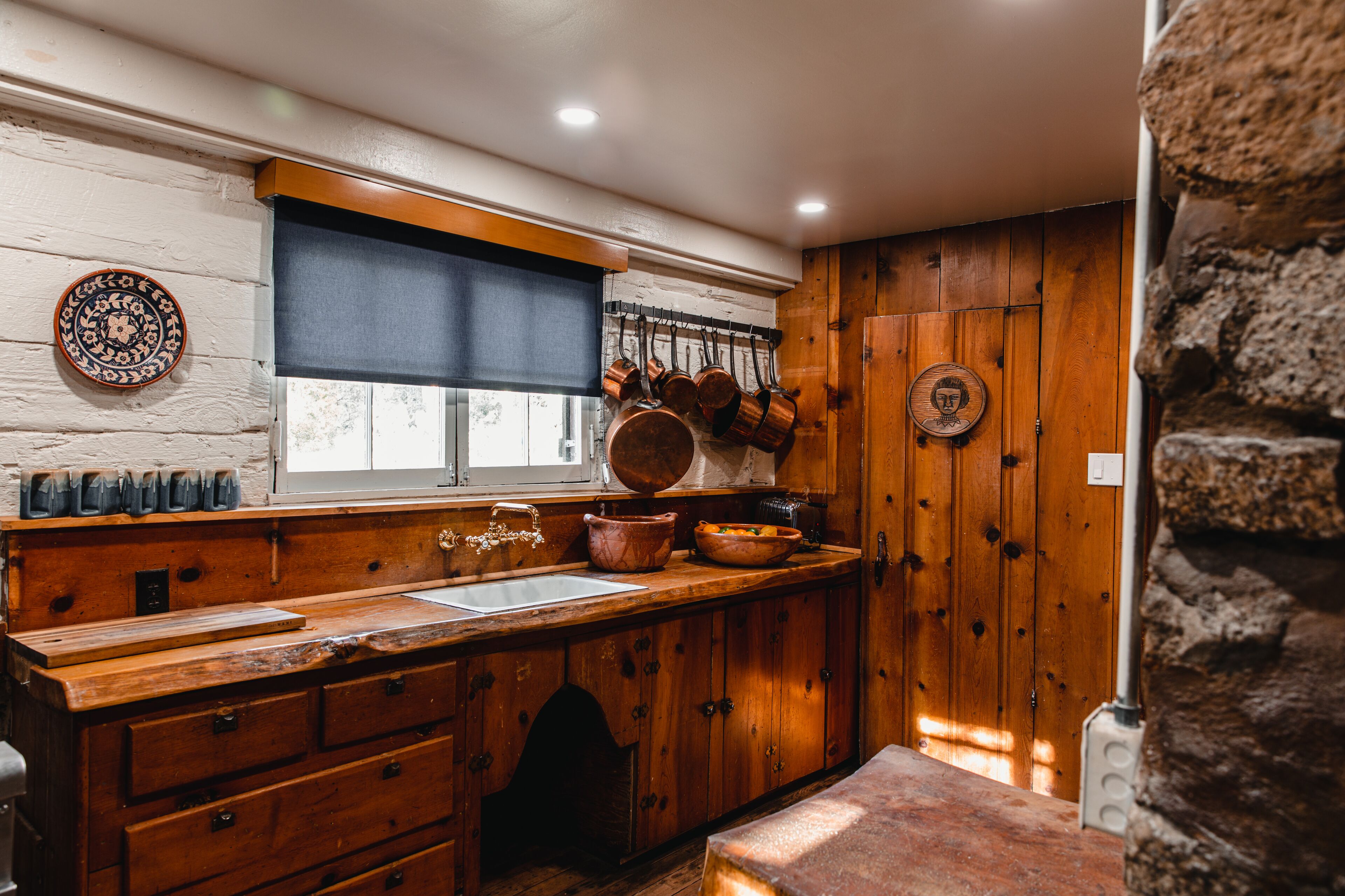 A rustic kitchen with wooden cabinets, a large window with blinds, and hanging pots.