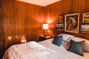 A wooden-paneled room with a bed, pillows, a nightstand, a lamp, and framed pictures on the wall.