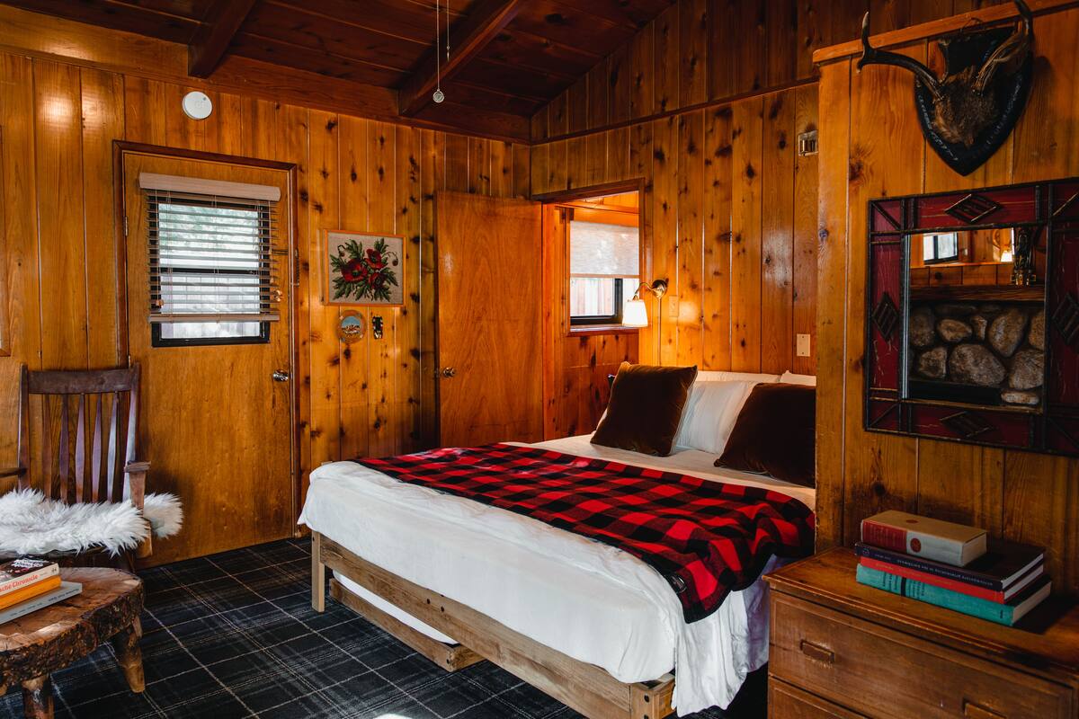 A wooden cabin bedroom with a bed, a chair, a small table, and a fireplace.