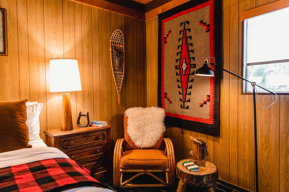 A wooden-paneled room with a bed, a chair with a fur throw, a wall hanging, and a lamp.