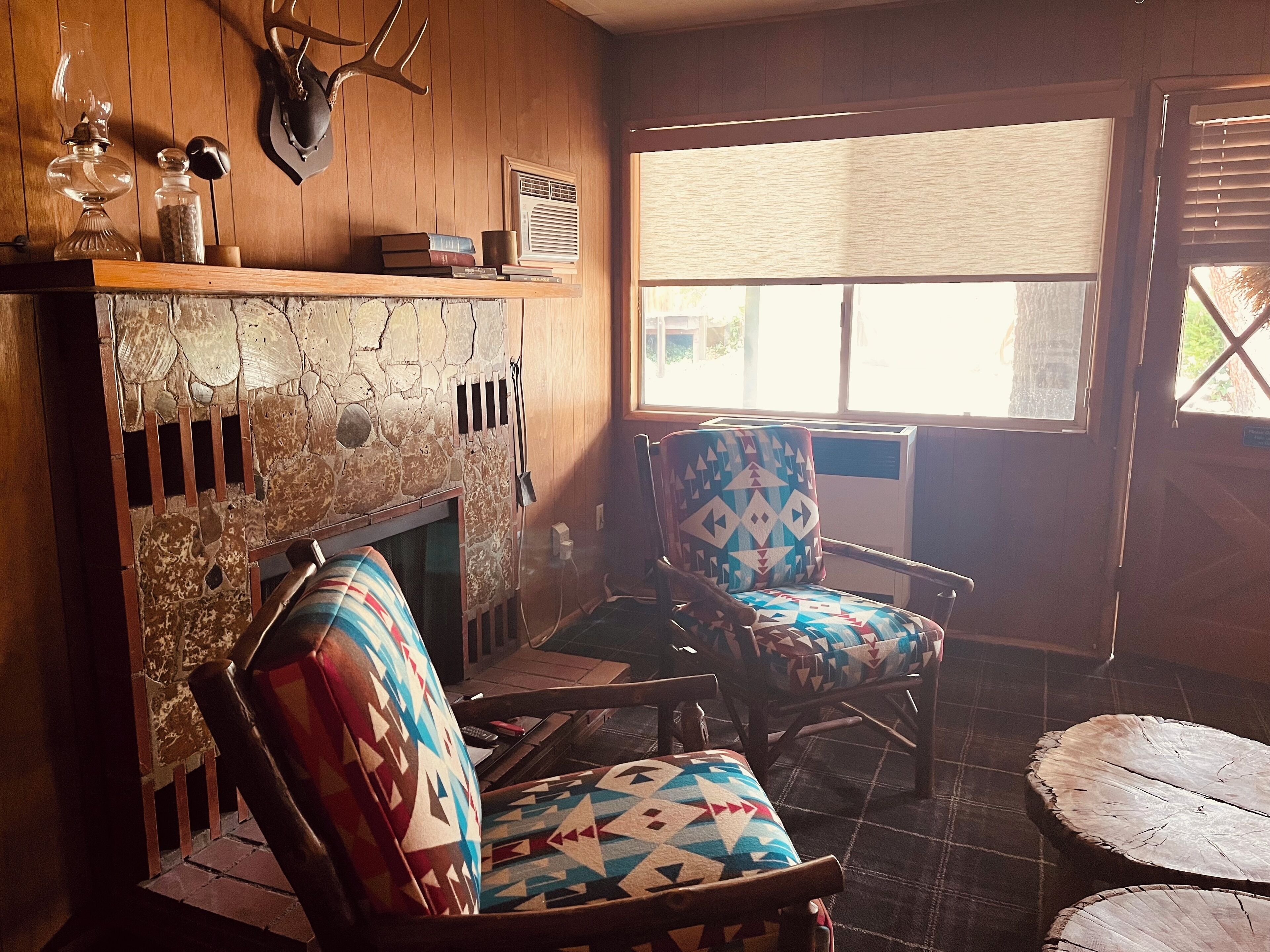A cozy room with a stone fireplace, two patterned armchairs, a wooden coffee table, and a window with blinds.
