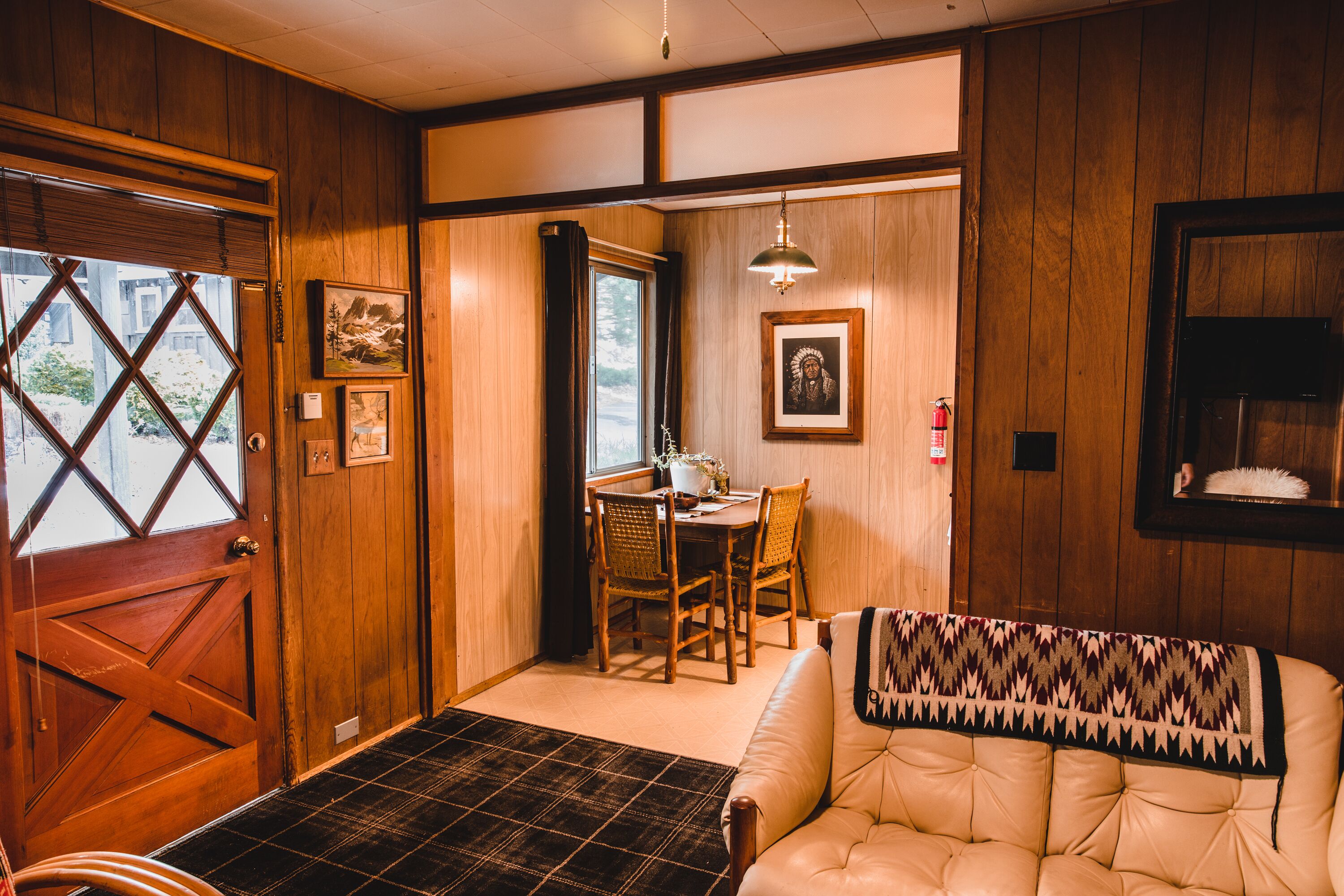 A room with a wooden door, a dining area with a table and chairs, a sofa, and a hanging light fixture.