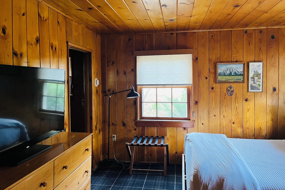 A wooden-paneled room with a bed, a desk, a television, a window with blinds, and two framed pictures on the wall.