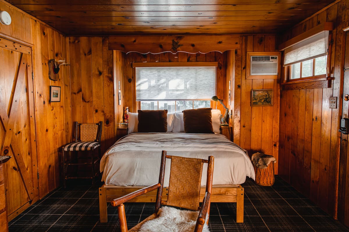 A wooden cabin bedroom with a bed, two chairs, a window, and a lamp.