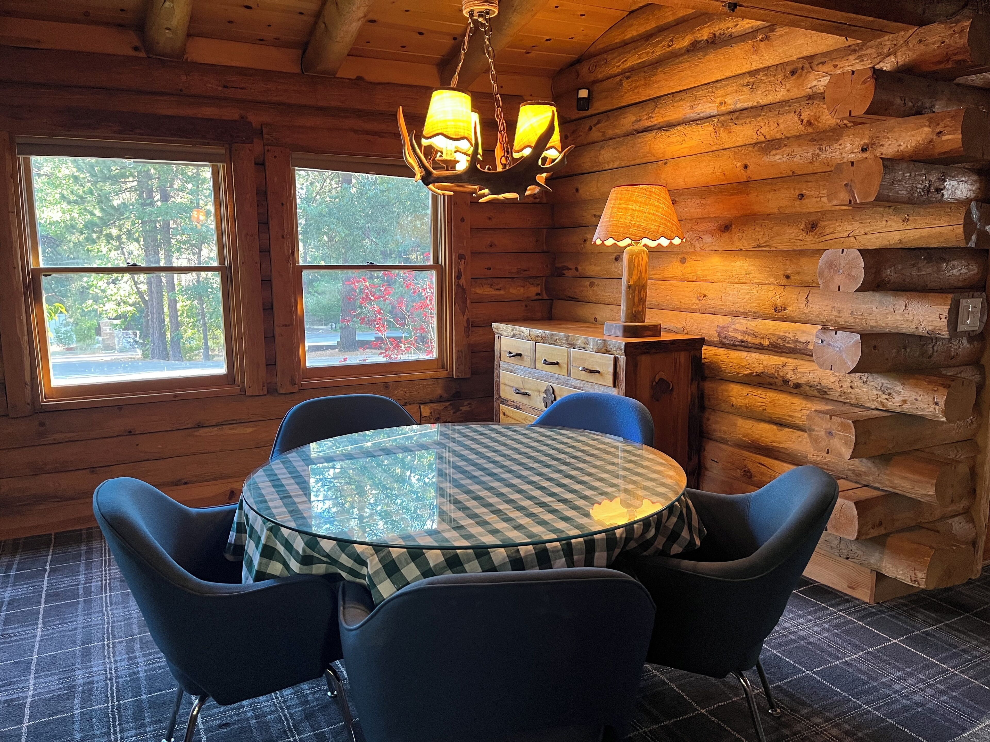 A wooden cabin interior with a round table and chairs, a window with a view of trees, and a lamp on a dresser.