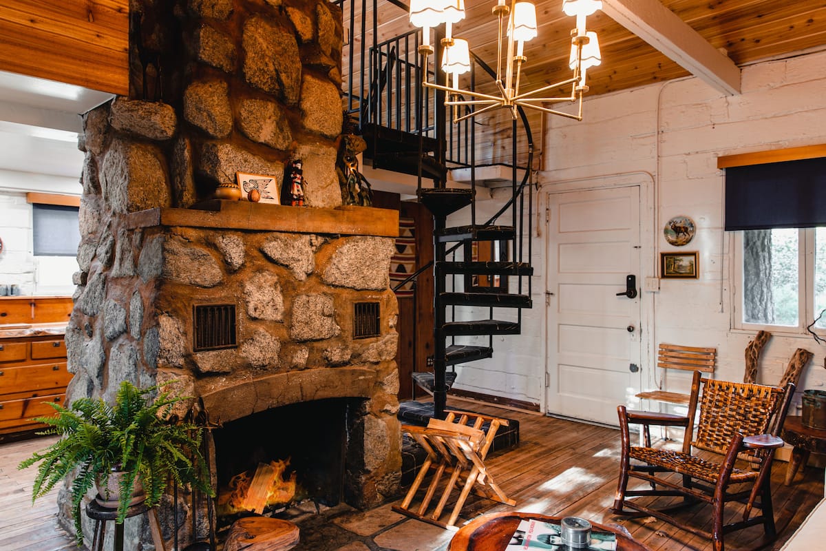 A rustic interior with a stone fireplace, wooden flooring, a spiral staircase, and a dining area with a table and chairs.