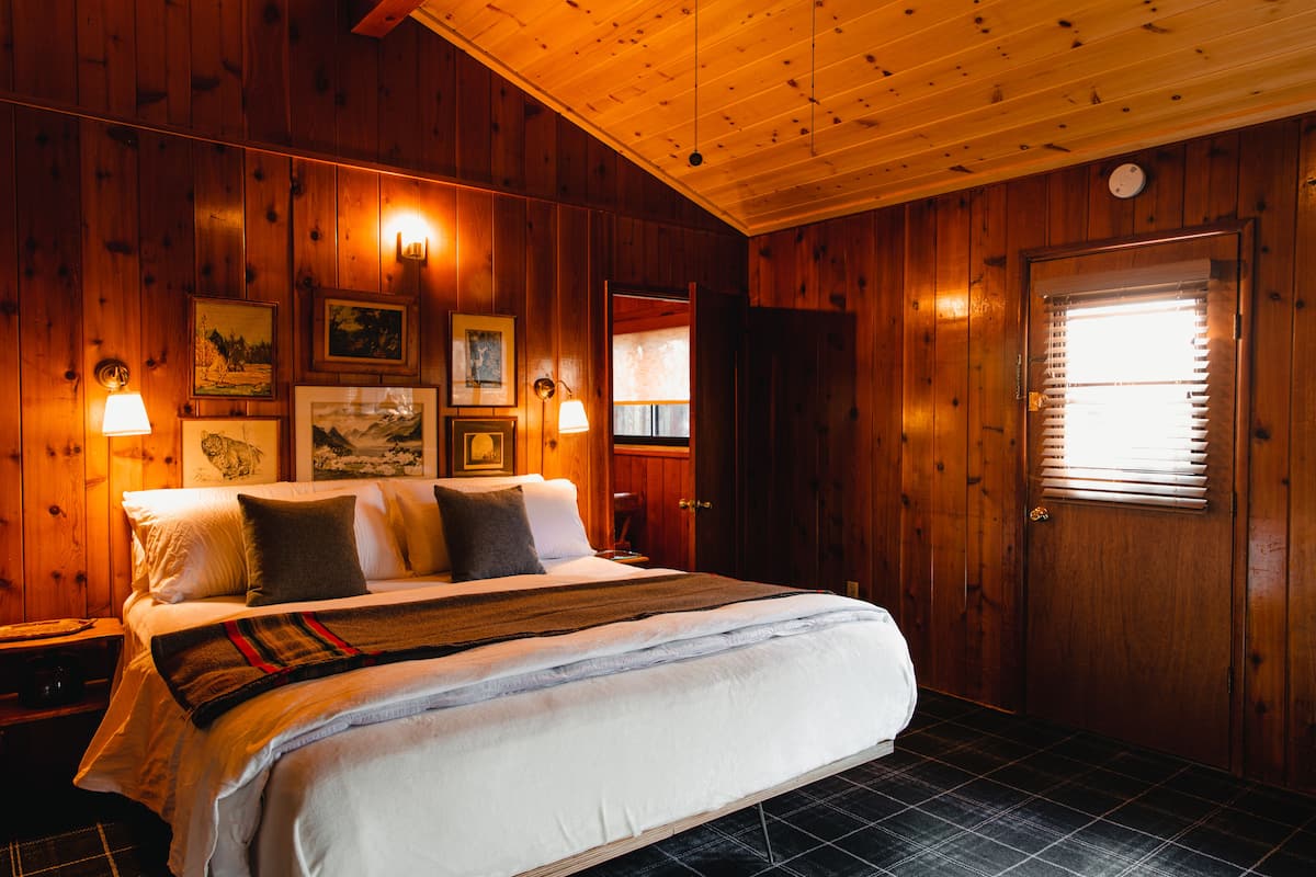 A wooden-paneled bedroom with a bed, framed pictures, and a window with blinds.