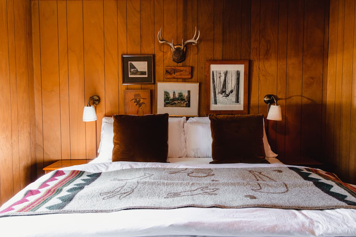 A wooden-paneled room with a bed, two pillows, a blanket, and framed pictures on the wall.