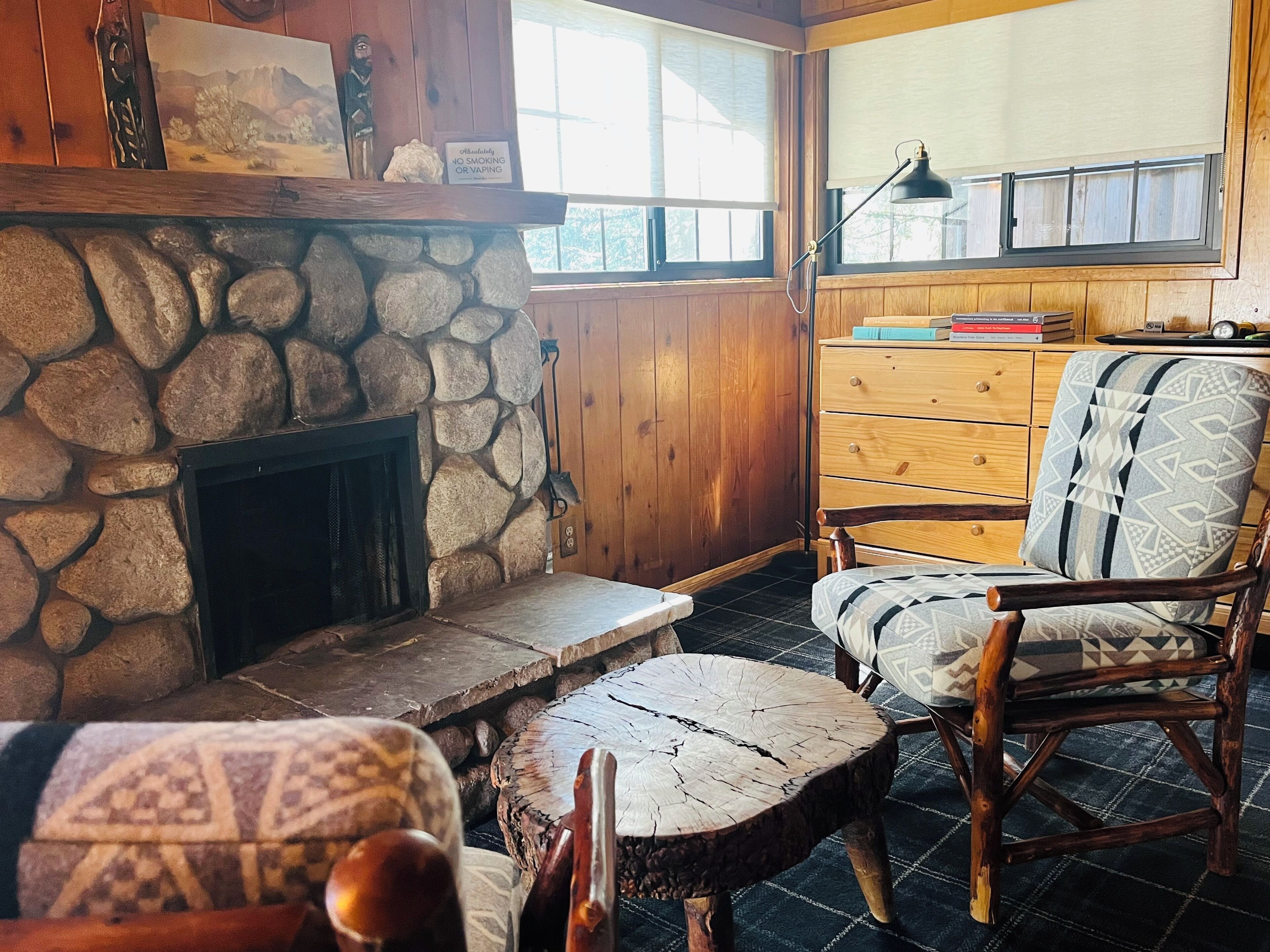 A cozy room with a stone fireplace, wooden walls, a wooden dresser, and a patterned armchair.