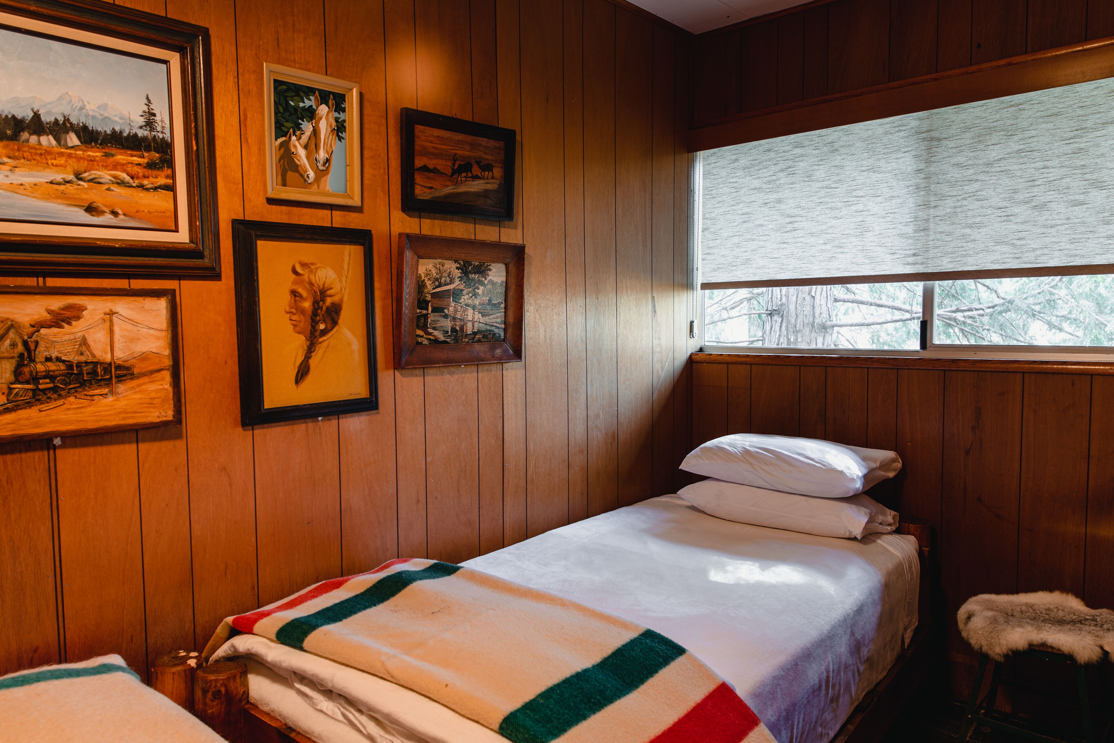 A wooden-paneled room with a bed, a window with blinds, and several framed pictures on the wall.