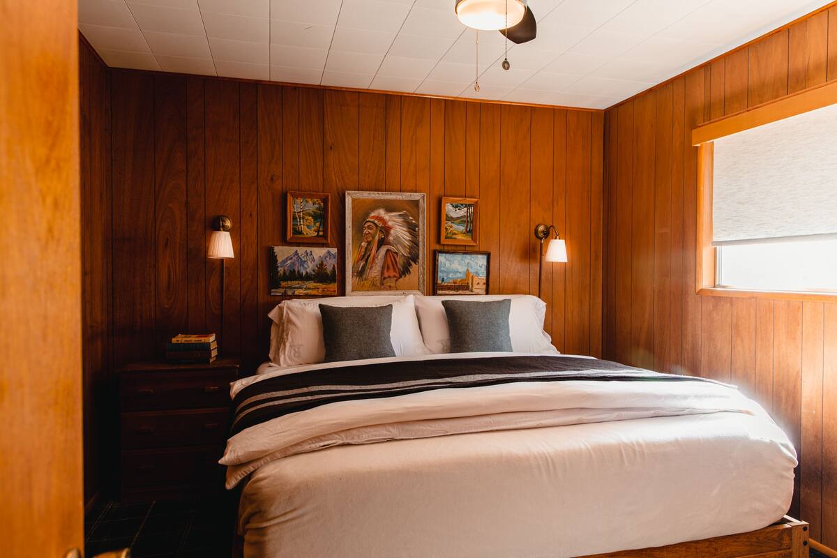 A wooden-paneled room with a bed, two wall-mounted lamps, and framed artwork on the wall.