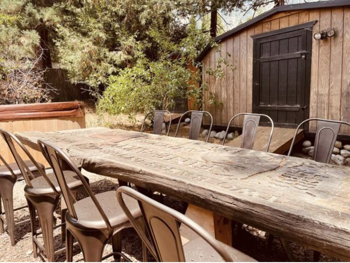 A rustic wooden table with metal chairs, surrounded by a wooden fence and trees.