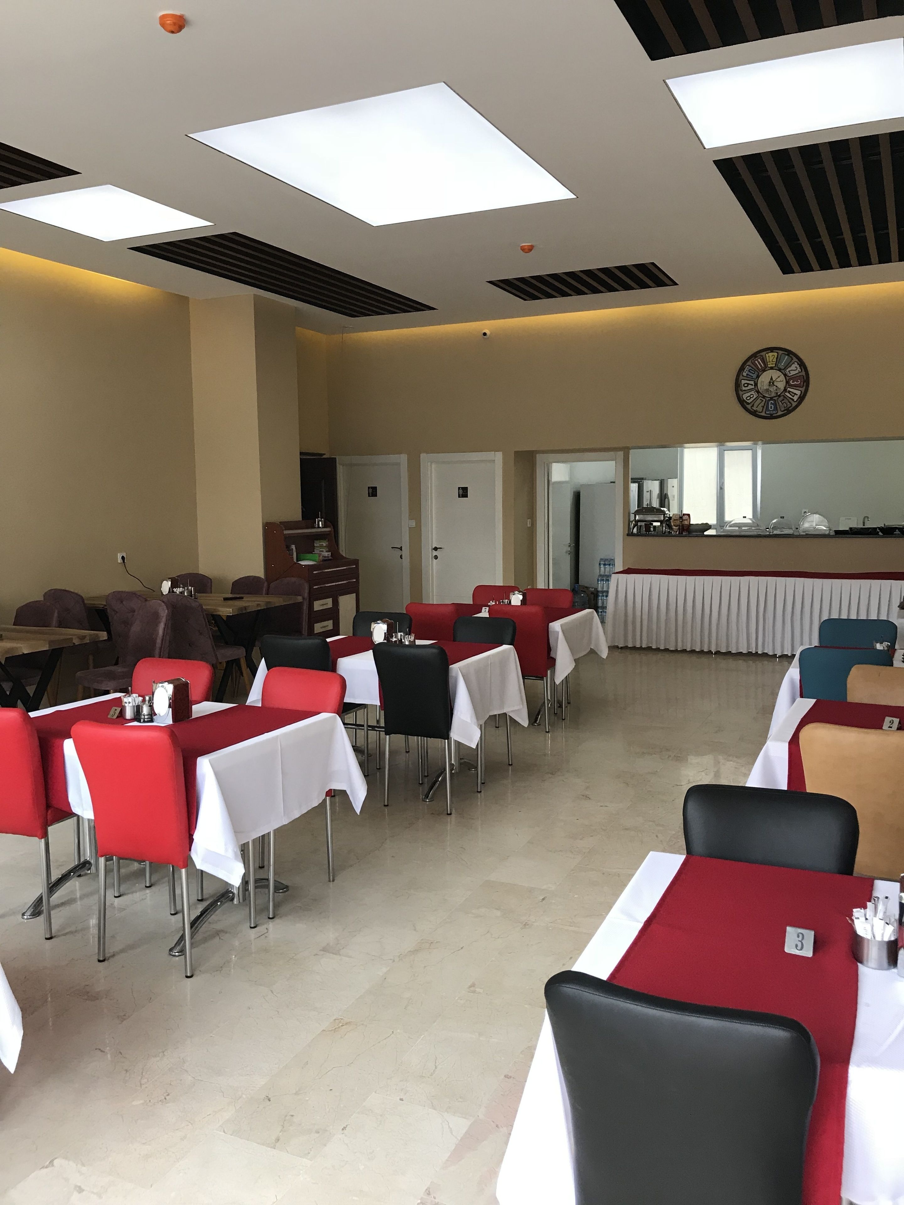 A dining area with tables and chairs, a clock on the wall, and a kitchen in the background.