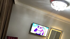 A hotel room with a TV displaying an animated character, a wooden desk, a red armchair, a white chair, and a mirror with lighting. - Ankara Atlantik Otel (Ankara)