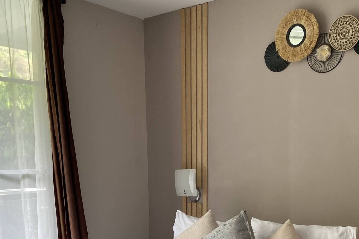 Un lit bien fait, avec une couette blanche, des oreillers gris et beige, et un plaid marron. Un panneau en bois orne le mur derrière le lit, et un luminaire mural est fixé au-dessus.