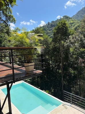 Une piscine dotée d’une terrasse en bois et de rampes en métal, entourée d’une végétation luxuriante et avec une chaîne de montagnes en arrière-plan.