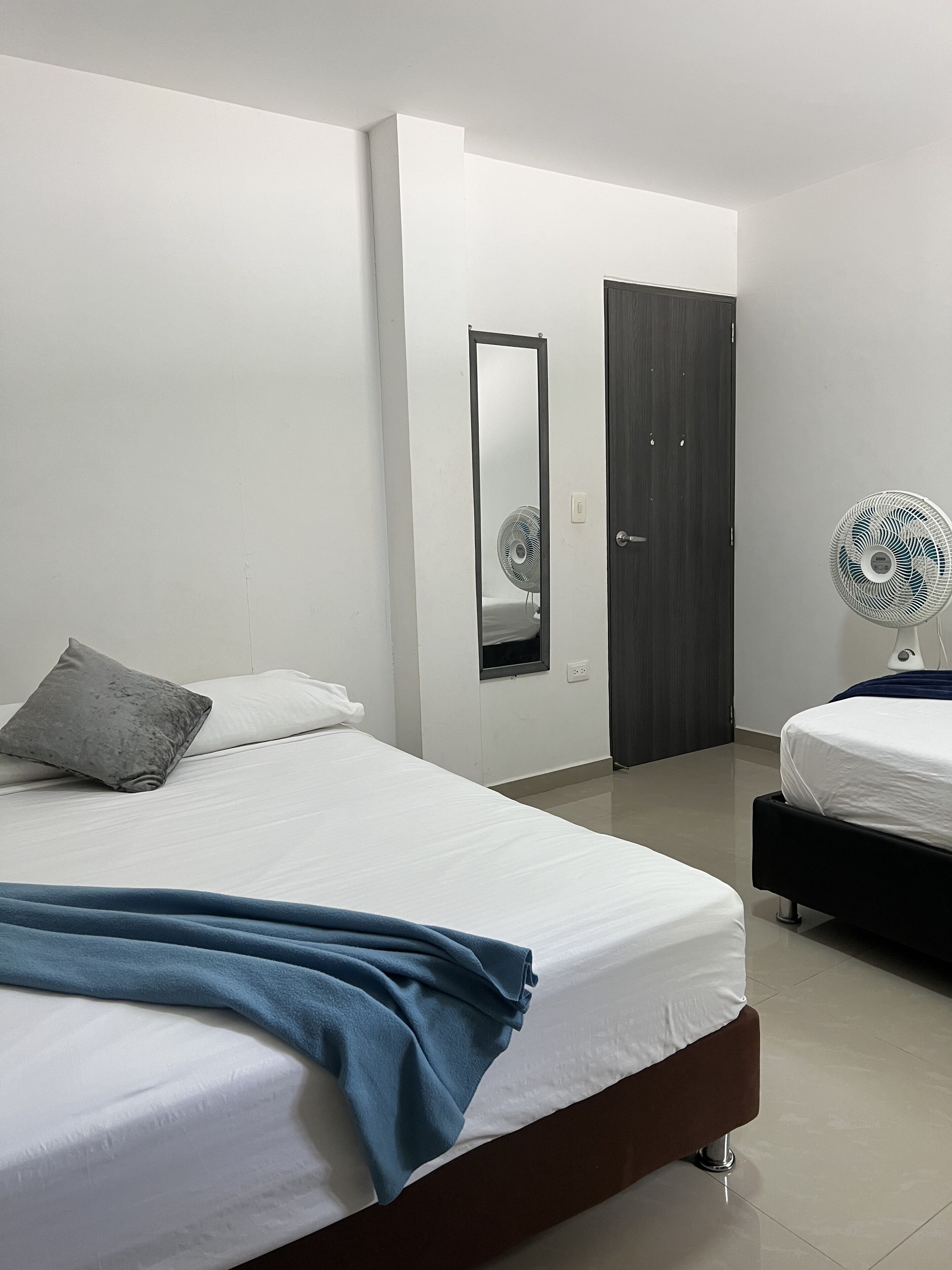 A modern bedroom with a large bed, a bedside table, a mirror, a wardrobe, and a standing fan.