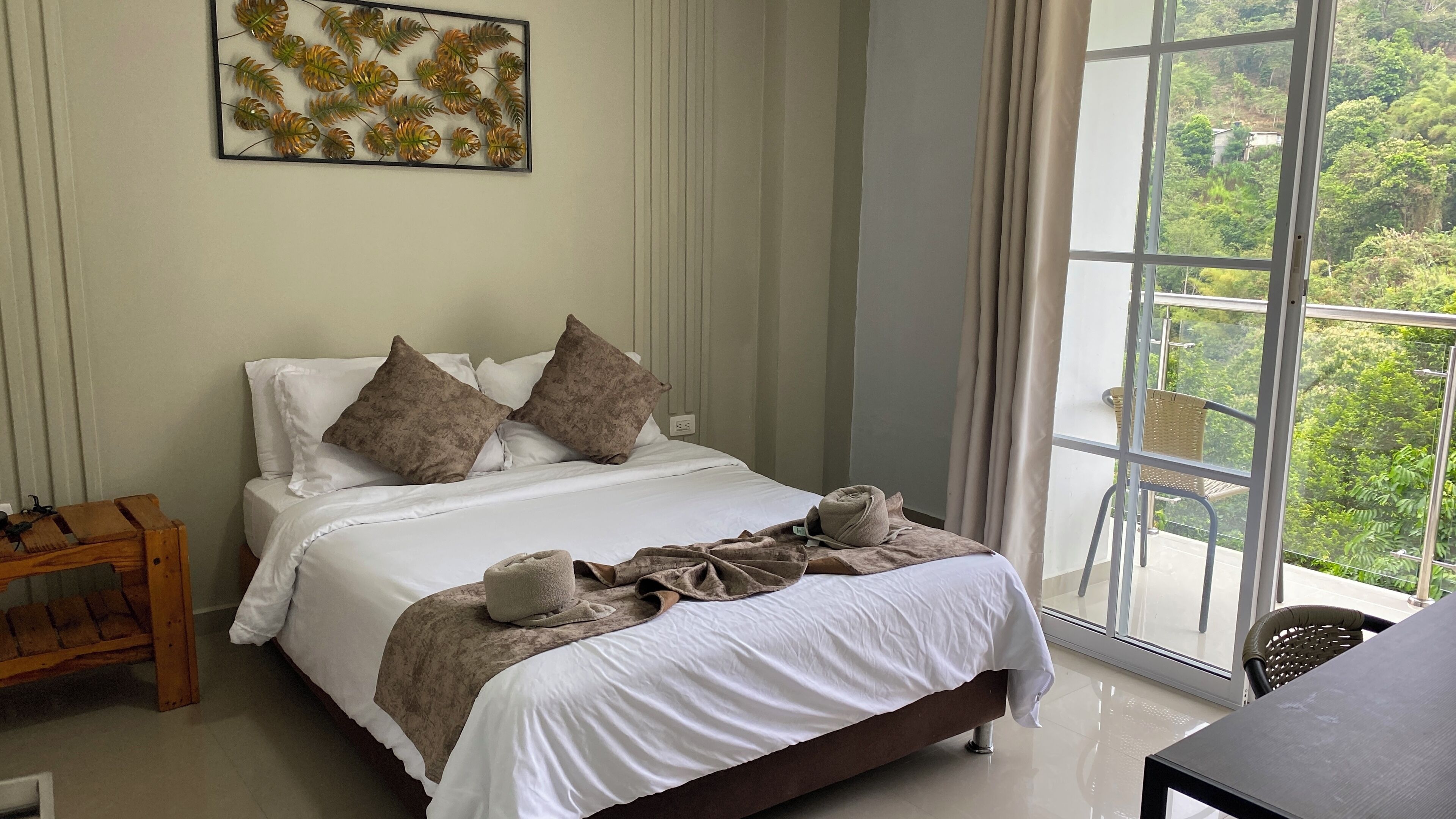 A bedroom with a bed, pillows, a blanket, a wooden nightstand, and a window with a view of greenery.