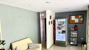 Un espace d’accueil comprenant un canapé, un réfrigérateur rempli de boissons, un tableau d’affichage et une plante en pot.